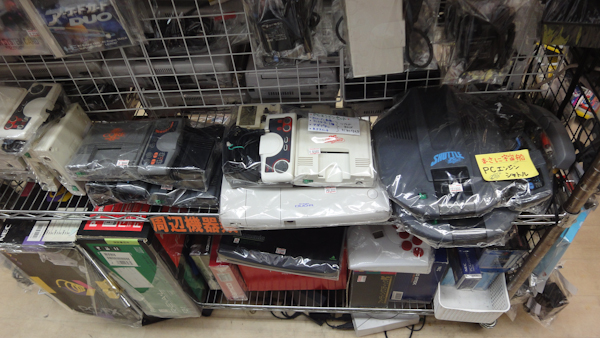 retro video game consoles on a shelf, mostly pc engine variants